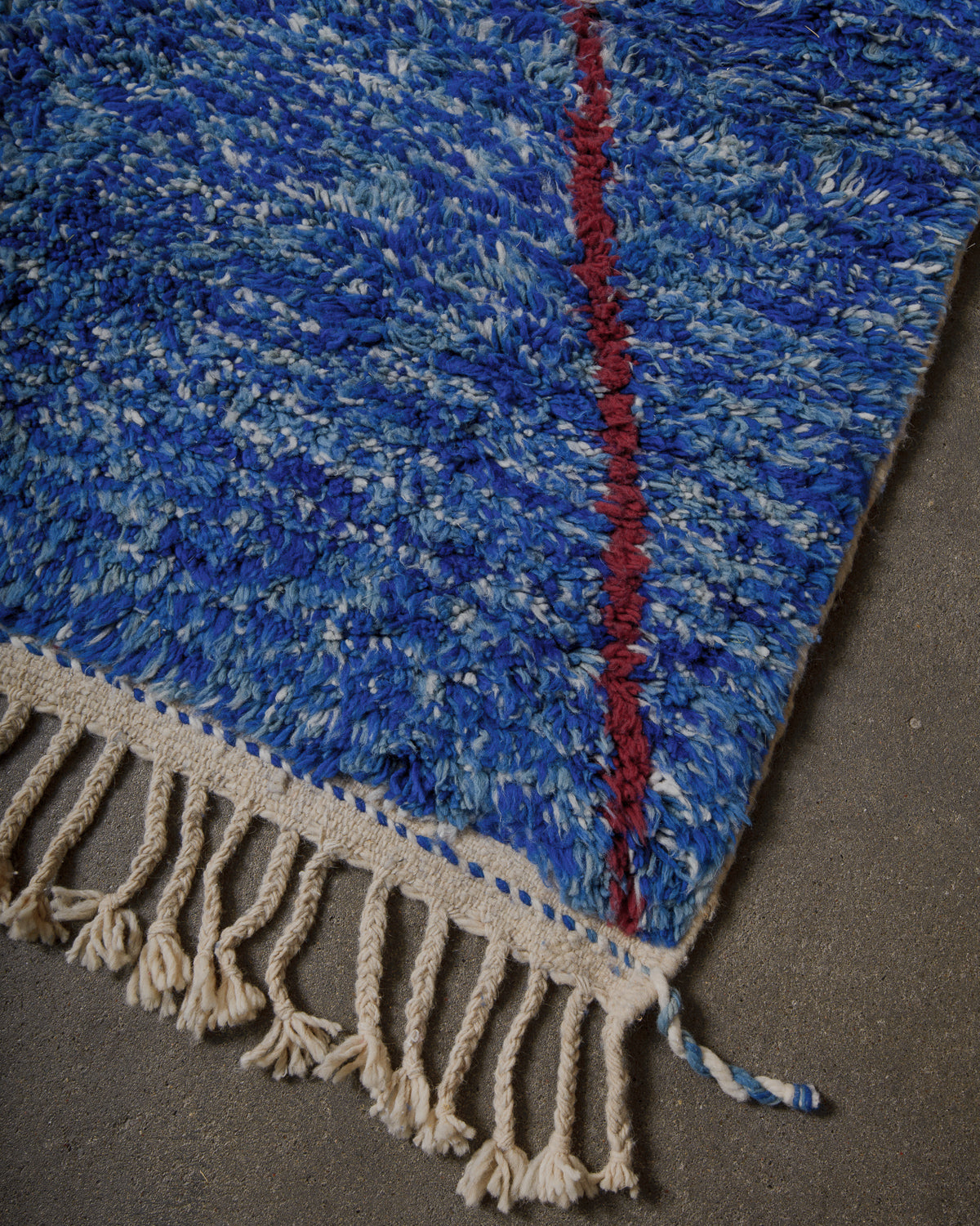 Corner of blue wool rug with natural wool tassels on concrete floor.